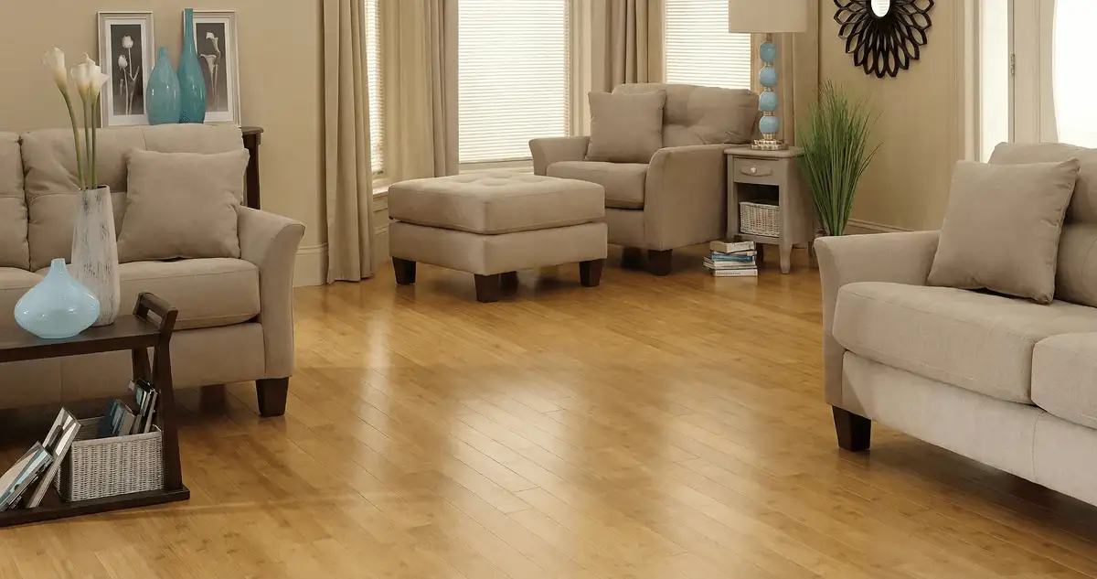 A living room with professionally installed bamboo flooring, beige sofas, a matching ottoman, a side table with a blue lamp, vases, framed pictures, and large windows with blinds and curtains in Knoxville, TN